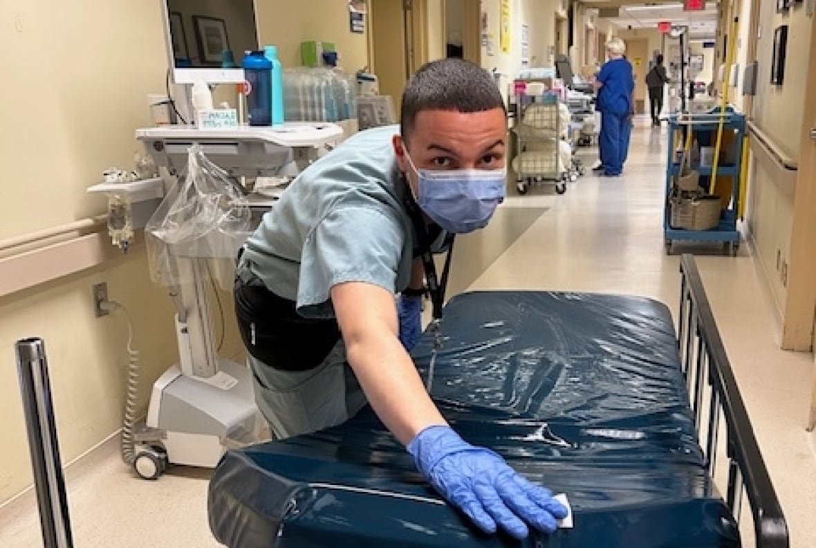 A young man in a surgical mask, gloves and medical scrubs wipes down a transport bed in a hospital hallway.