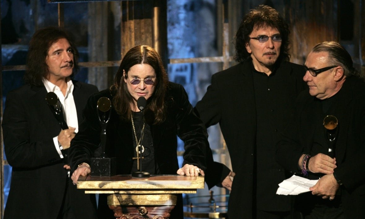 Four men wearing black stand together. The second from the left is leaning down to speak into a microphone on a podium, his hands braced on the podium. 