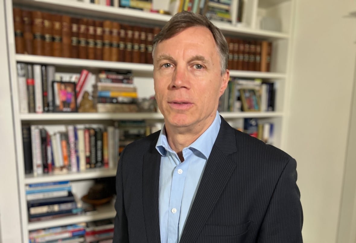 A man with silver hair wears a blue button-down shirt and dark jacket as he stands in front of an office bookcase. 