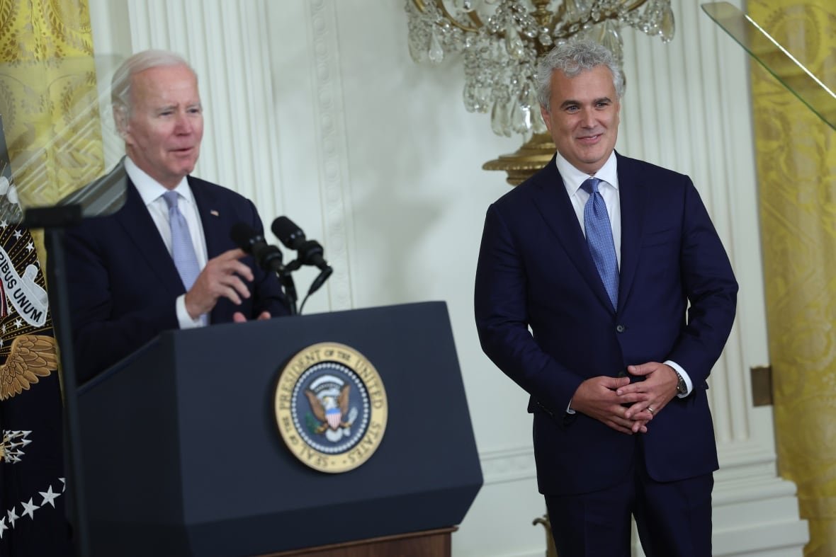 Two cleanshaven, white haired men stand in an ornate room. One of the men is behind a podium.