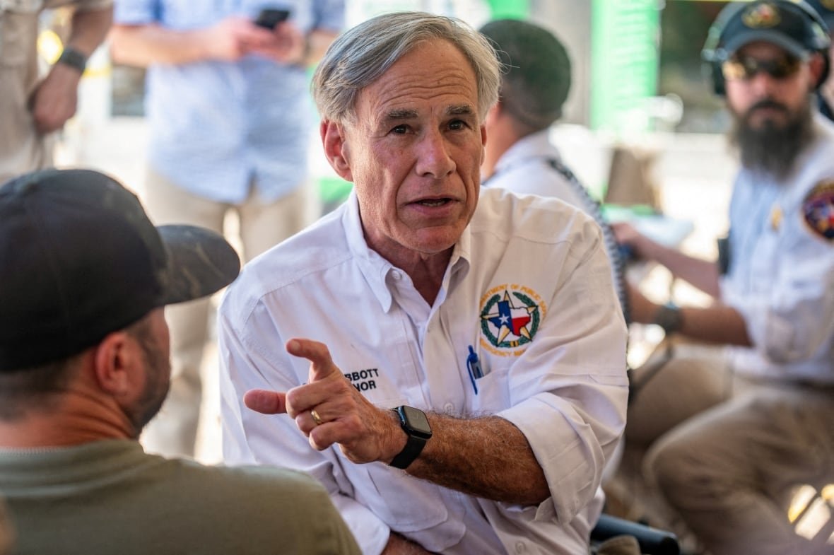 Grey haired man in a white shirt with a Texas state logo on the chest gestures as he speaks