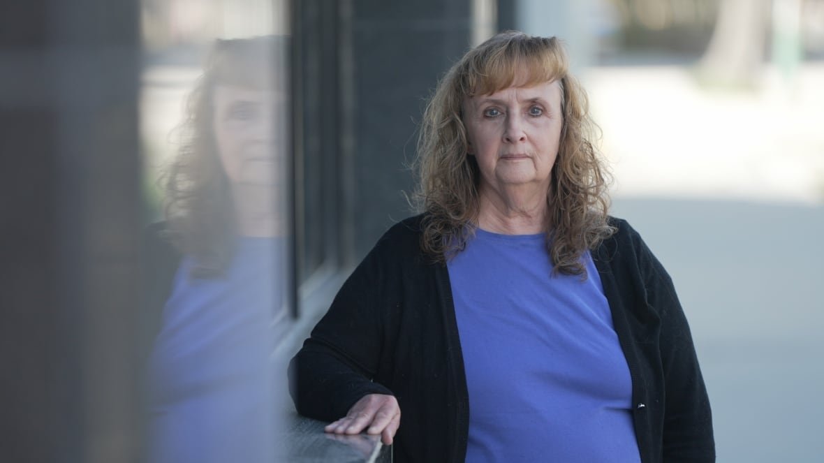 A woman wearing a blue shirt and black sweater stands outside leaning against a windowsill.