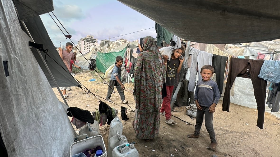 A woman stands with her back to the camera as several children and a man carry bags into a tent.