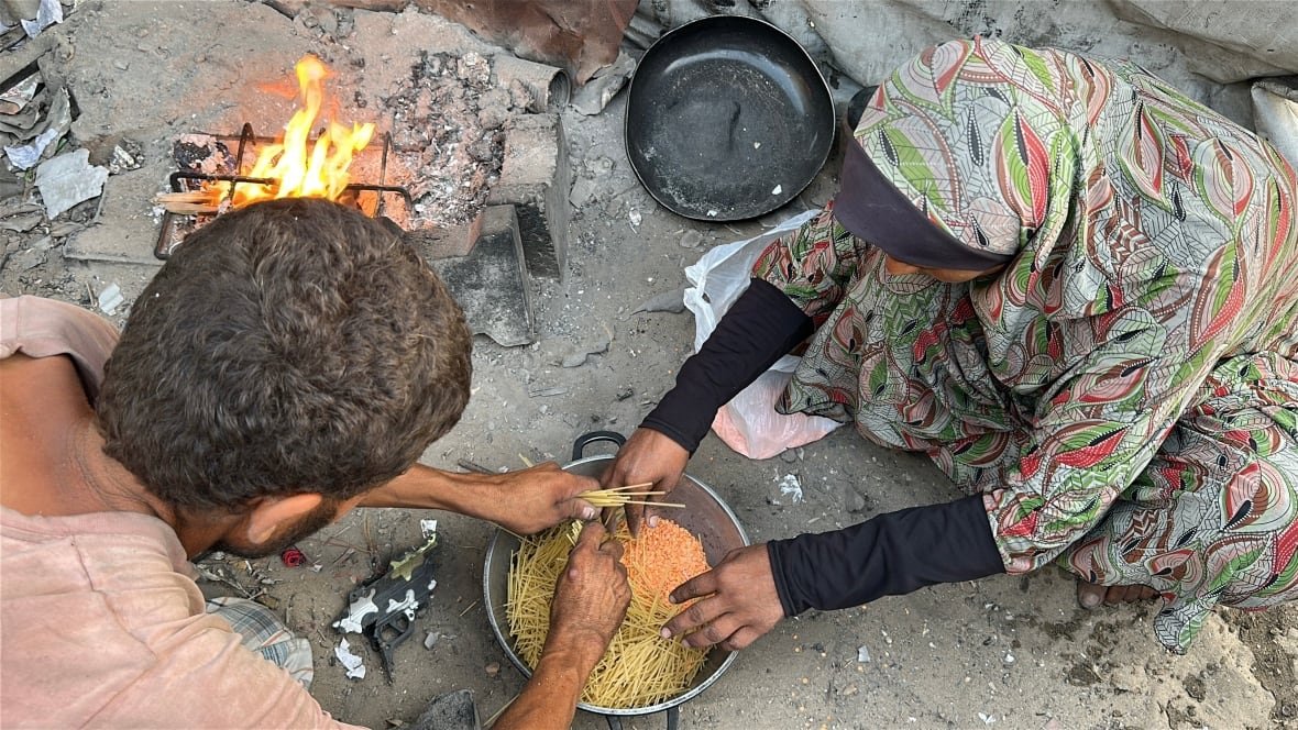 A man in a faded t-shirt and a woman in a hijab bend their heads over a pot containing uncooked spaghetti and red lentils as a fire burns at their side.