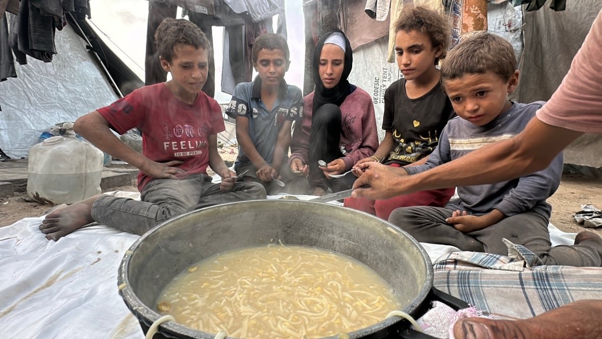 Five children sit on a blanket on the ground, watching as a man dishes out noodles from a steaming pot.