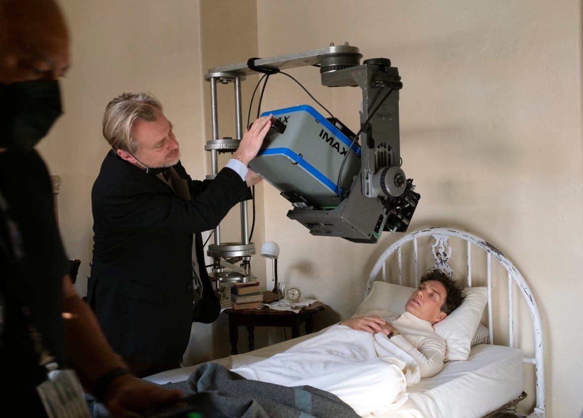 A blond man in a dark suit jacket adjusts an Imax camera hanging over a thin man in a bed. 