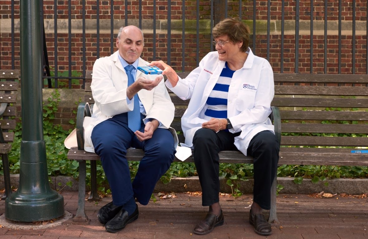 Dr. Weissman and Dr. Kariko are on what looks like a park bench outside of a building. She's giving him something out of a small box into his hand. He's looking very amused and she's laughing.