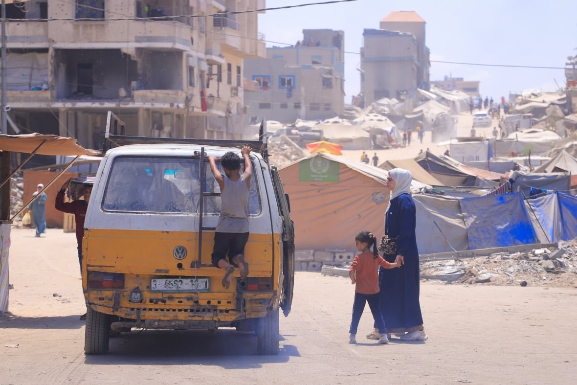 A woman standing with a child looks toward a yellow and white van, with a young boy hanging off the back, as it stops on a dusty road. In the background are tents and partially destroyed buildings.