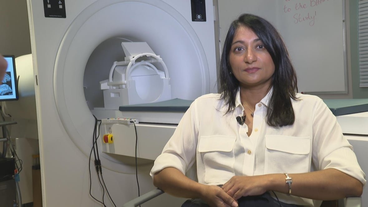 A woman with straight black hair stares at the camera with a slight smile. She is wearing a white blouse and a silver watch. There's an MRI machine behind her.