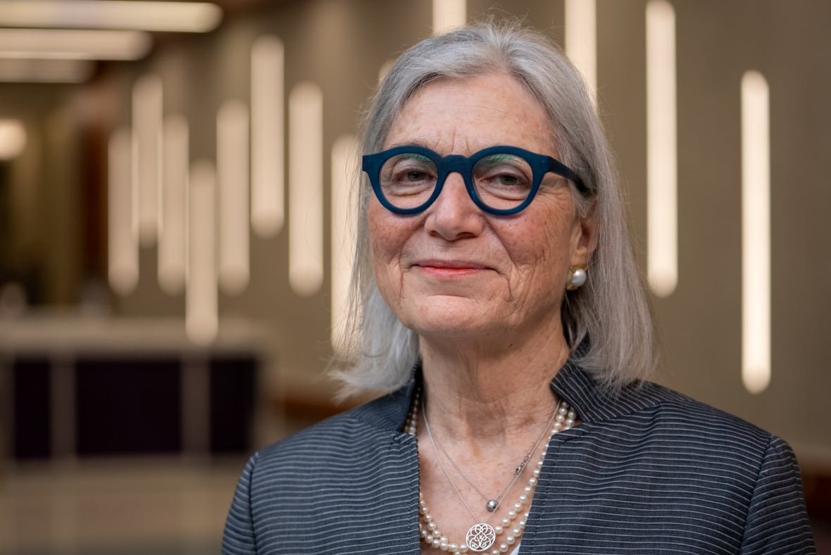 A woman with a grey bob and thick blue glasses smiles at the camera. She is wearing pearls and a grey-black blazer