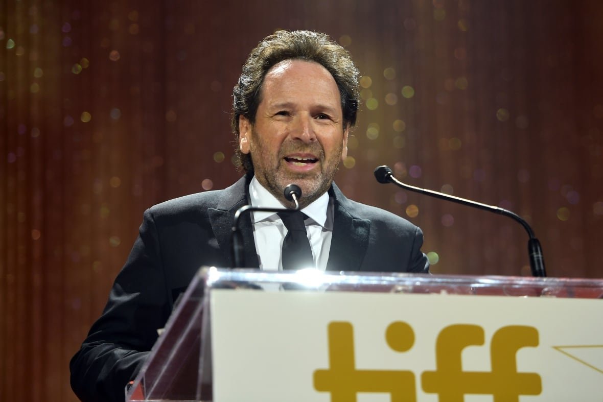 A man in a black suit stands behind a podium with the word "TIFF" on the front. the man is smiling while talking into the microphone on the podium and delivering a speech