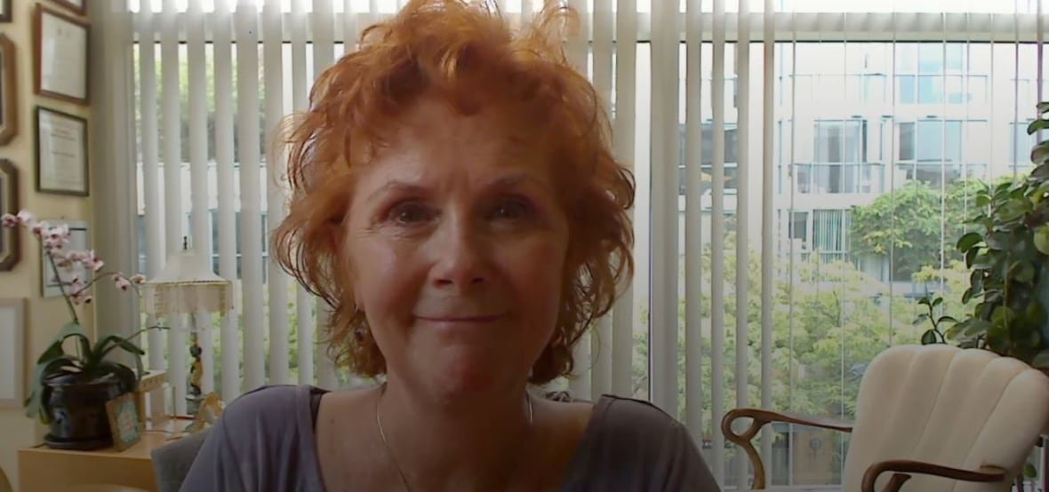 A woman with curly red, short hair smiles with white blinds, pink flowers and a white upholstered chair behind her.