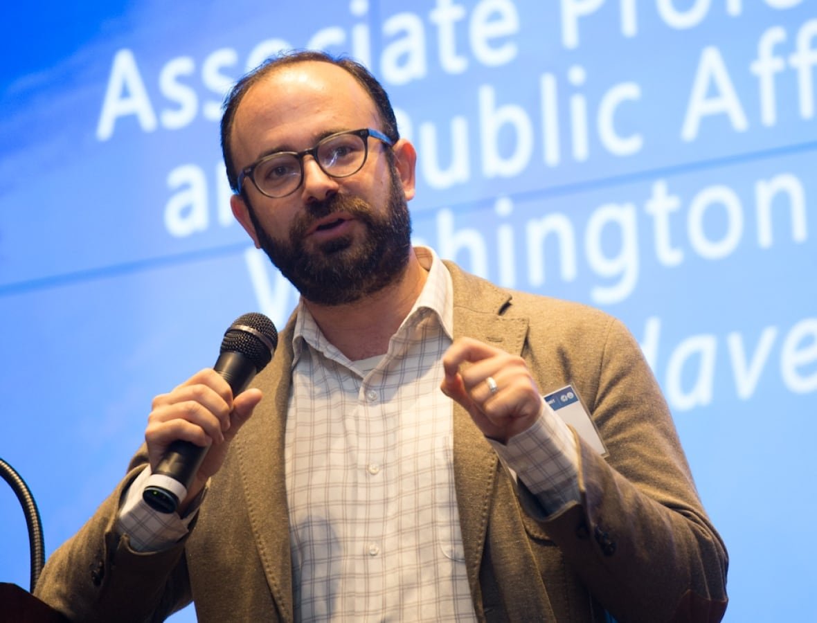 A man with glasses and a beard speaks on stage with a microphone.