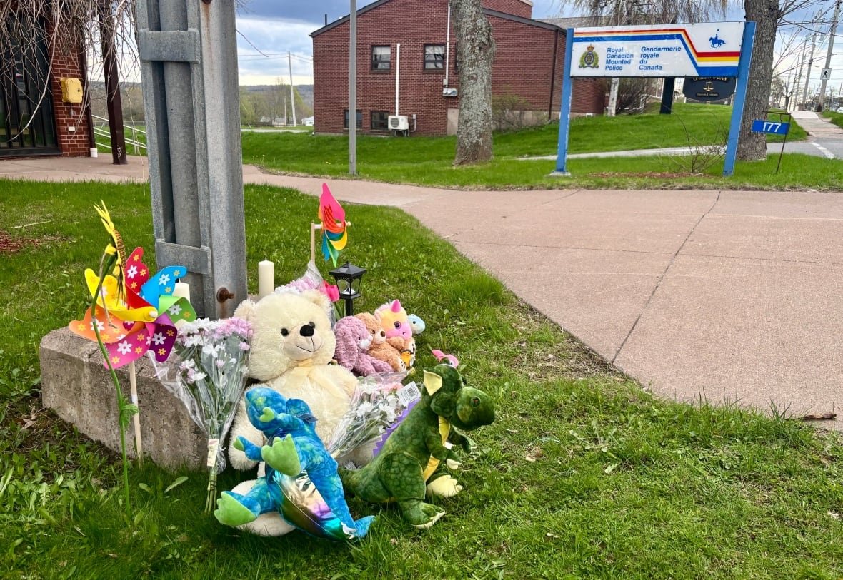A teddy bear and flowers are laid against a post outside an RCMP detachment.