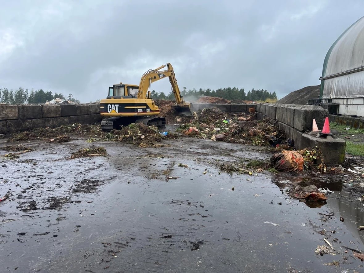 A yellow excavator scoops brown compost waste under a cloudy sky.