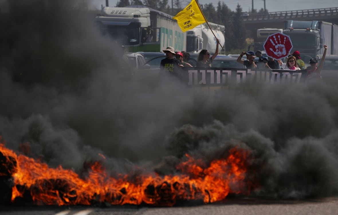 A thin line of fire at the bottom of the image crosses a road. Most of the image is obscured by smoke, but a group of people waving signs and flags is visible near the top, positioned behind the fire. 