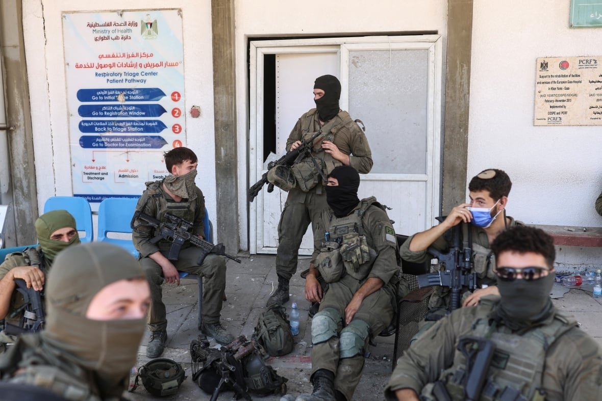 Soldiers, including several wearing balaclavas or masks, gather near the doors of a hospital building. One of the signs on the wall reads, 'Respiratory Triage Center'