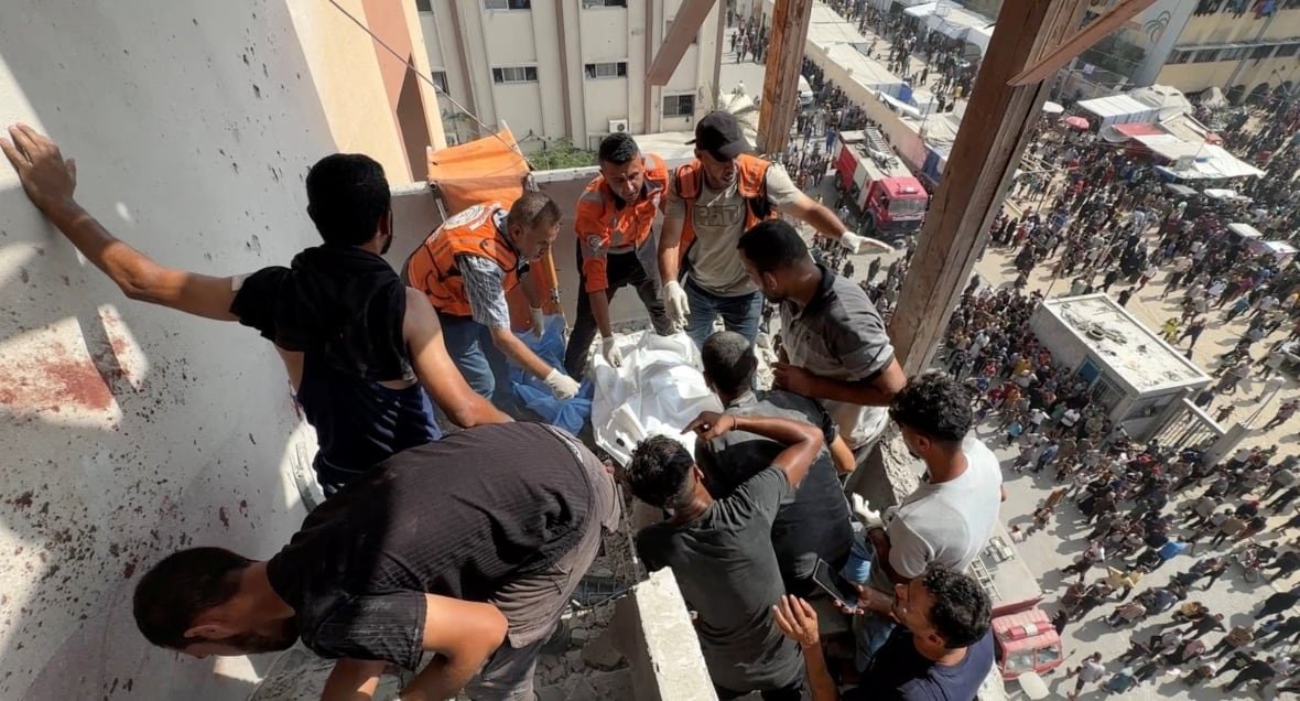 People, some of whom are wearing orange visibility vests, gather around what appears to be a body under a white sheet. 