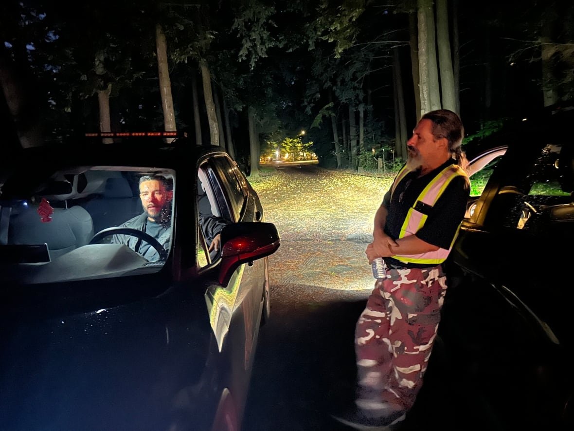 A man in a vehicle speaks with a man outside leaning against a second vehicle. It is evening. The man standing outside has a reflective vest on and wears his greying hair in a pony tail. 