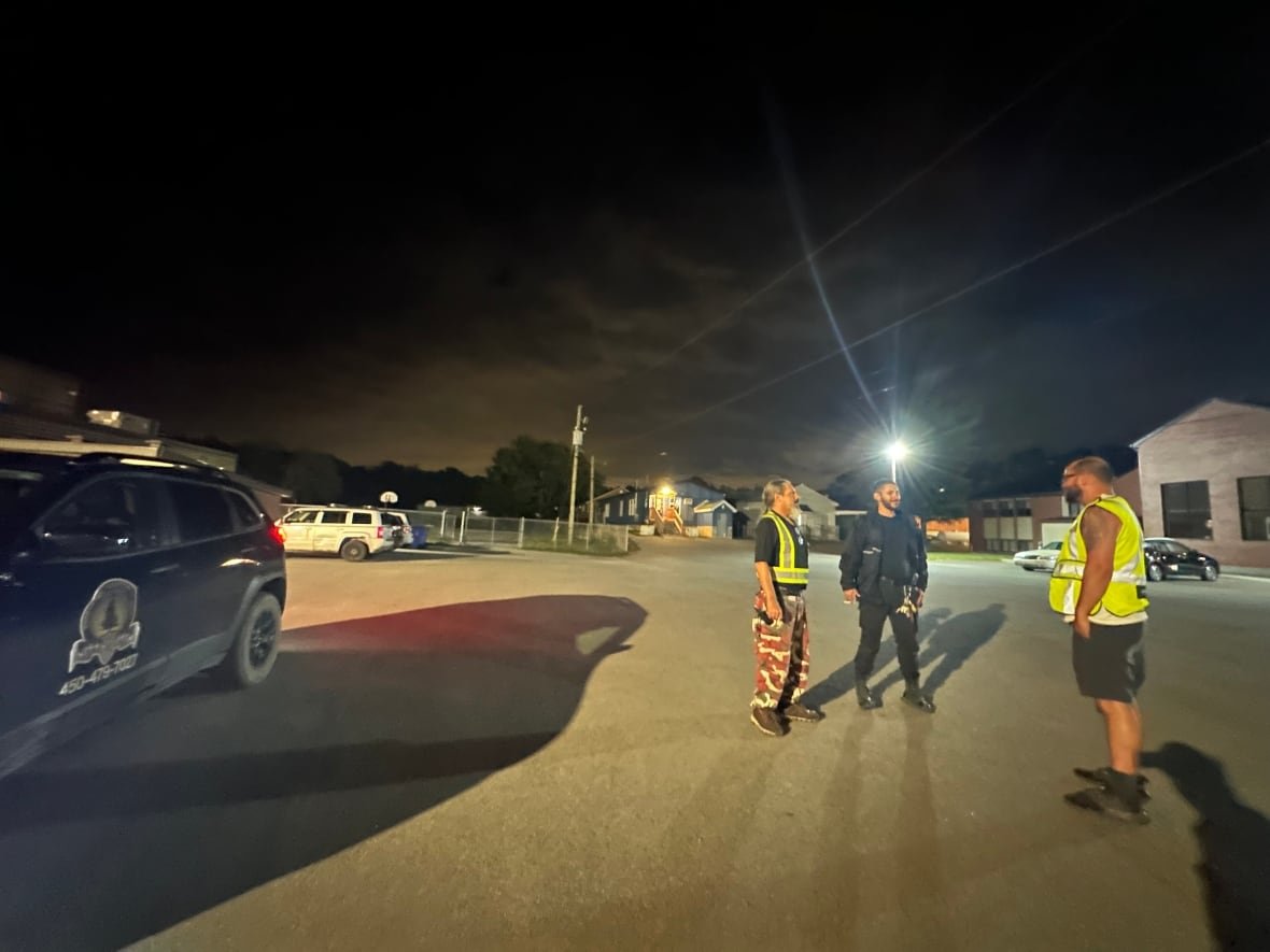 Three men stand in a parking lot at night to the right of the photo. A Kanesatake Perimeter Security Team SUV is parked to the left.