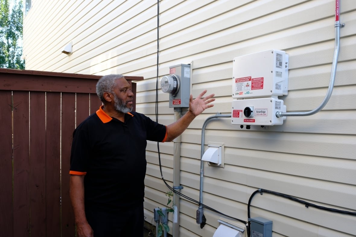 A man stands beside electricity meters and panels mounted on the side of his home. 