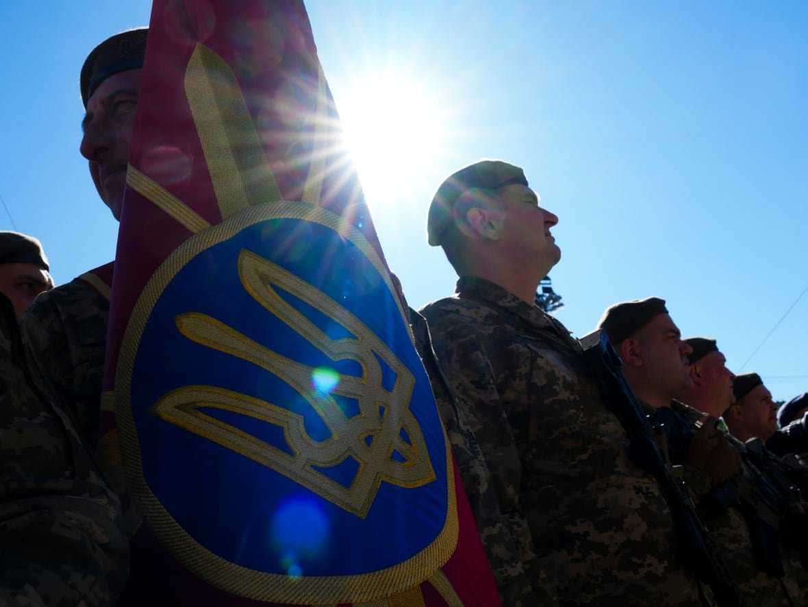 The sun blares behind a lineup of soldiers holding a Ukrainian ceremonial flag.