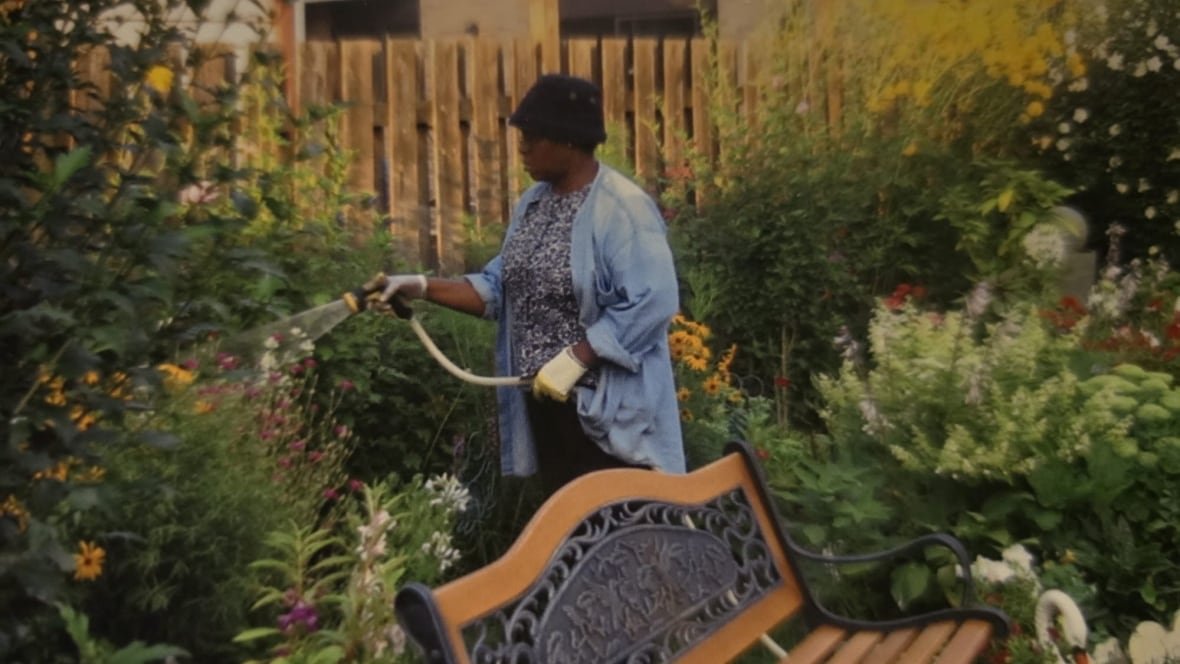 A woman wearing a blue garden coat waters a lush garden. She is wearing a black bucket hat.
