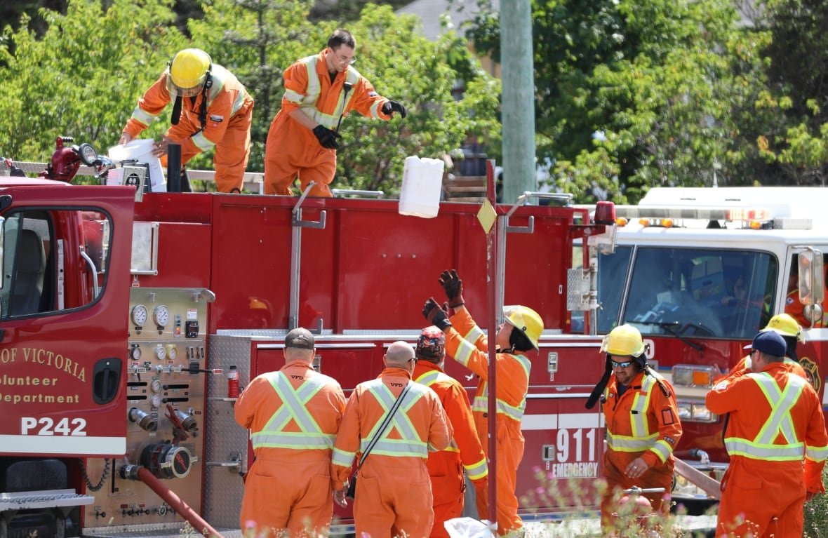 Man in orange suit standing on fire truck tosses town white box to group of men in orange suits