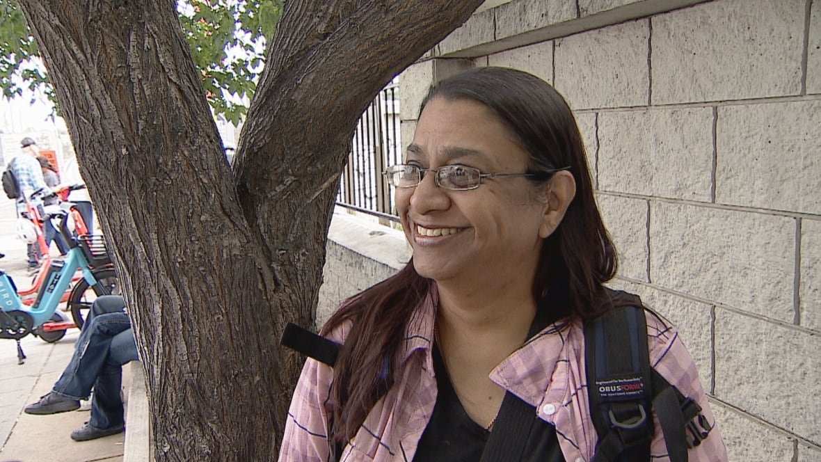 A woman wearing a backpack stands in front of a tree in the shade.