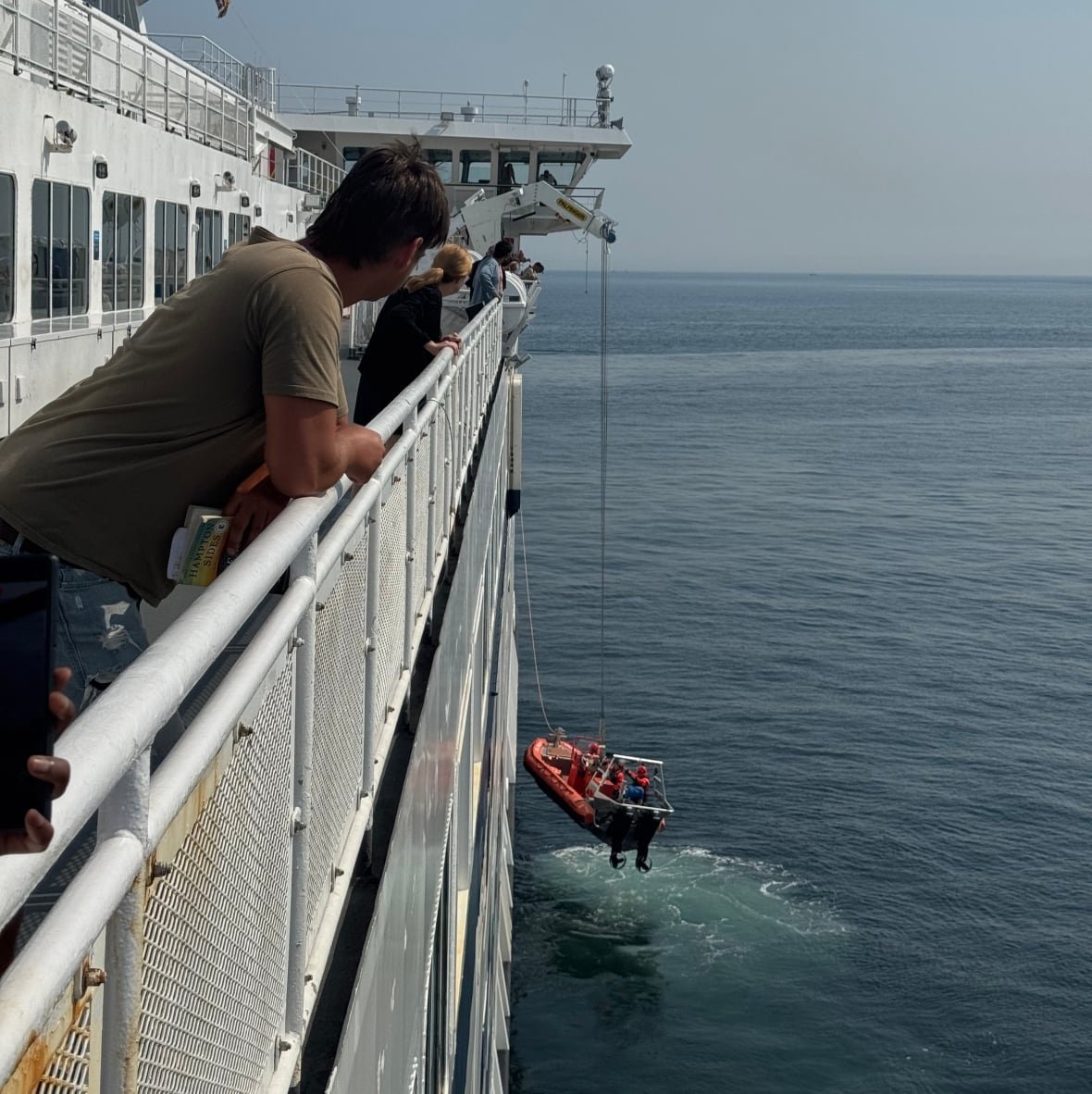 People are seen along a vessel's side as they look at a rescue boat being lowered from it.