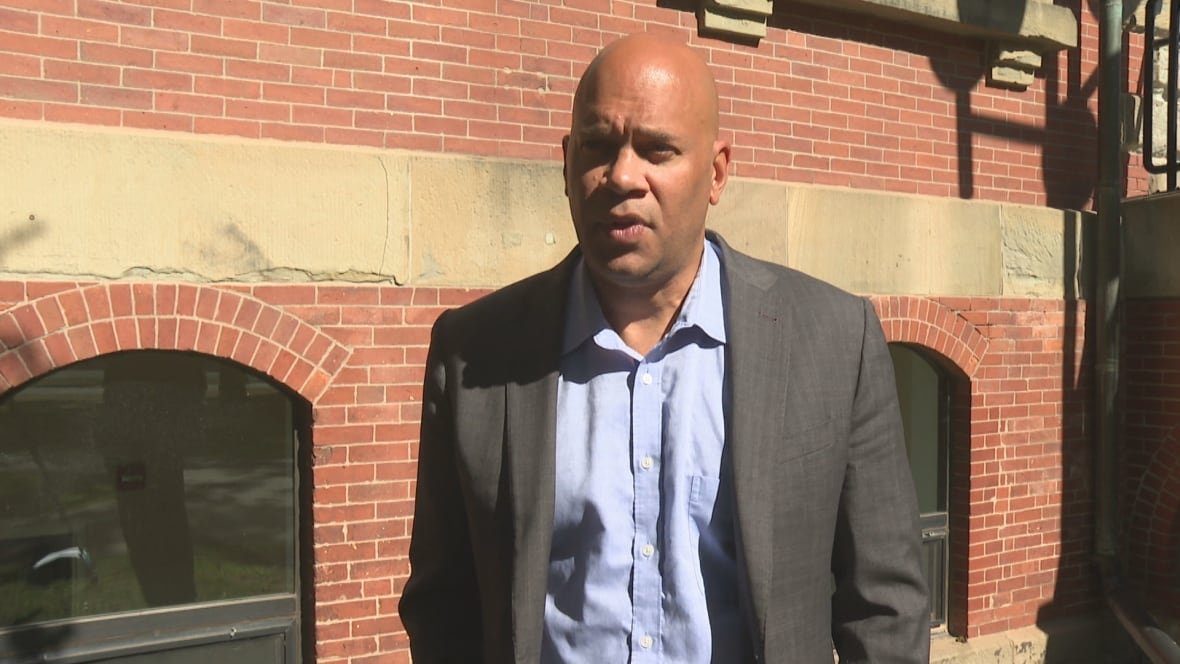 A bald man with a grey suit jacket over a pale blue button-down shirt stands in front of a brick building. 