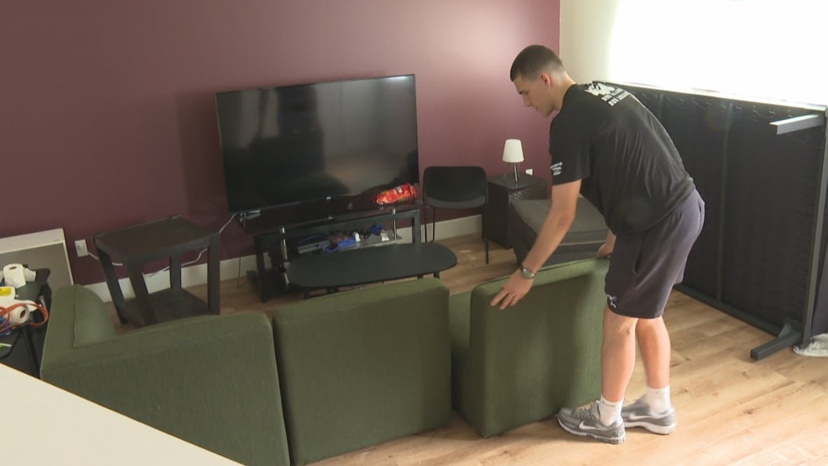 A young man in a black T-shirt and shorts pushes sections of a green couch together in front of a large TV set.