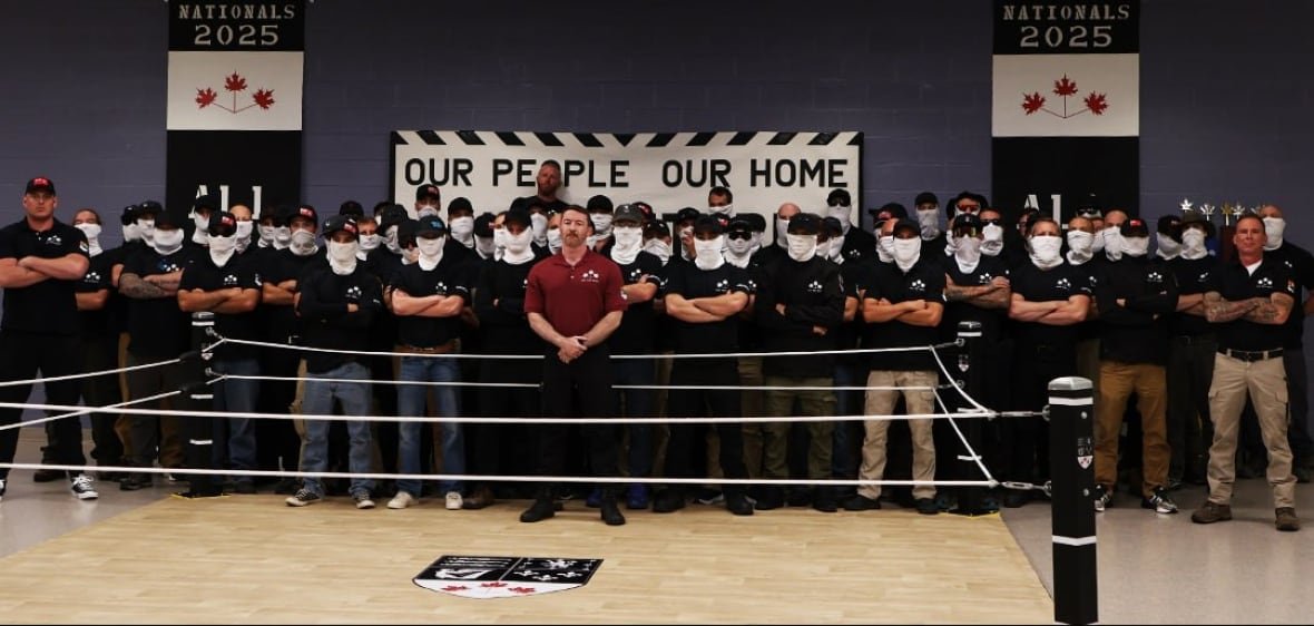 A group of mostly masked men in matching black T-shirts post behind a fight ring indoors.
