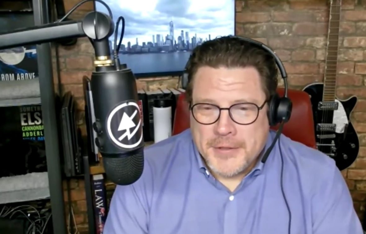 A man in a blue button up shirt and headphones sits in front of a microphone.