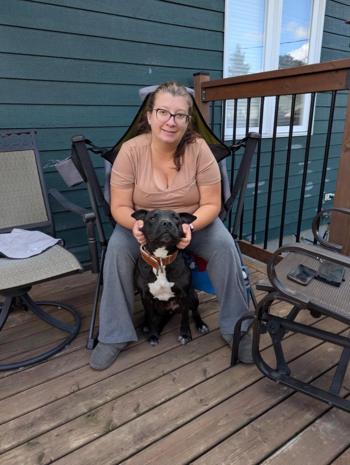 A woman wearing a beige t-shirt with grey pants sits on a deck, while petting a large black dog that sits between her legs.