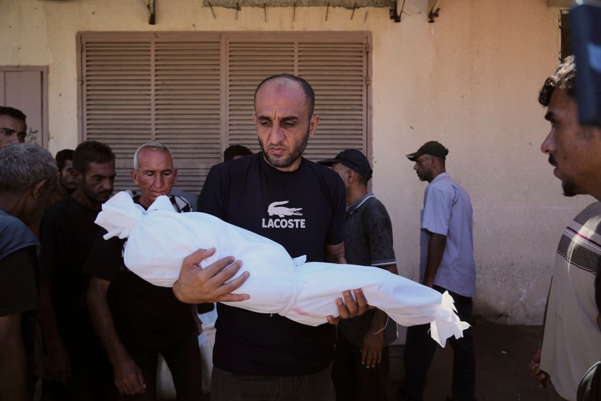 A man carries the body of a toddler wrapped in white material.