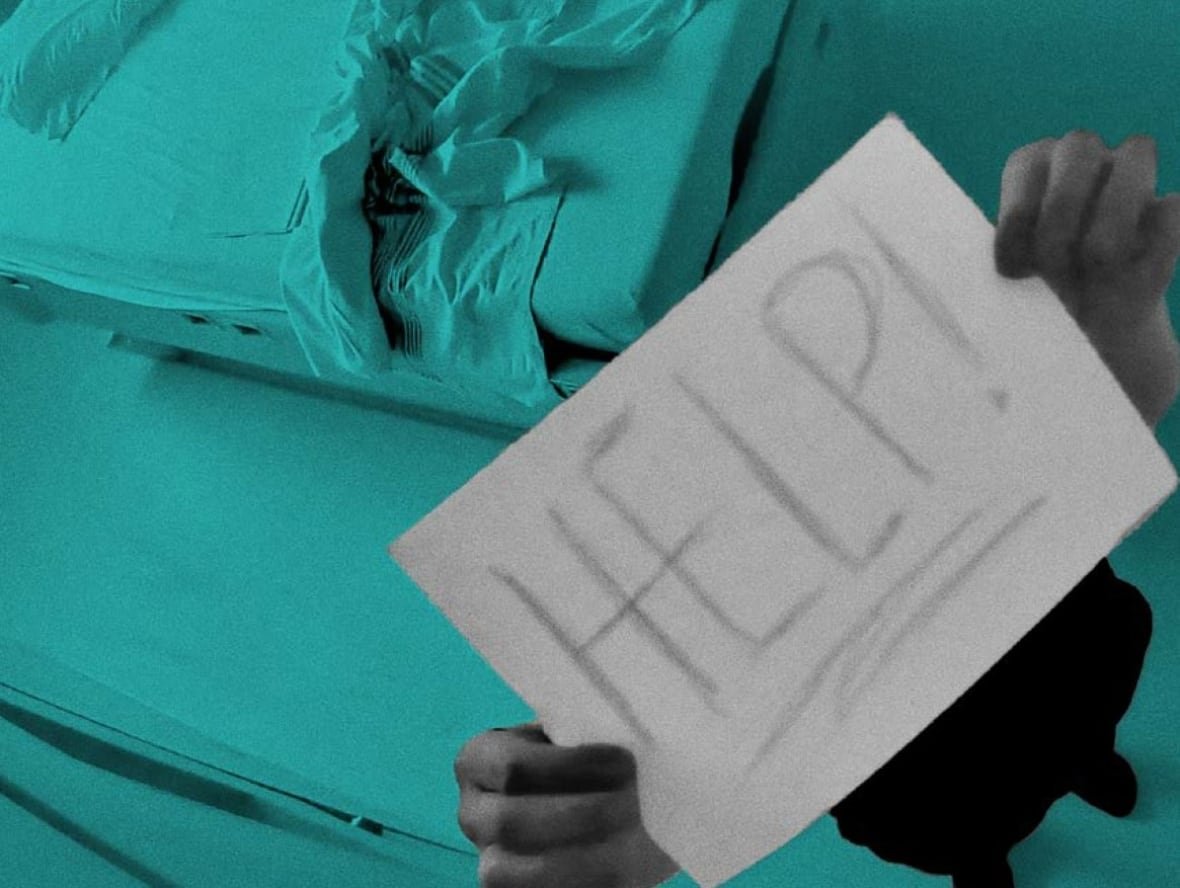 A person holds up a sign with the word "HELP!" handwritten. Their face is behind the sign. A bed with a sheet bunched up at the end and a pillow at the head is shown in the background.