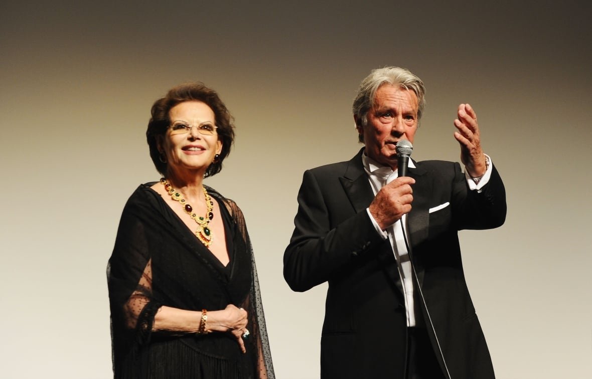 A dark haired woman and white haired cleanshaven men, both senior citizens, are both shown in formal wear and standing on a stage.