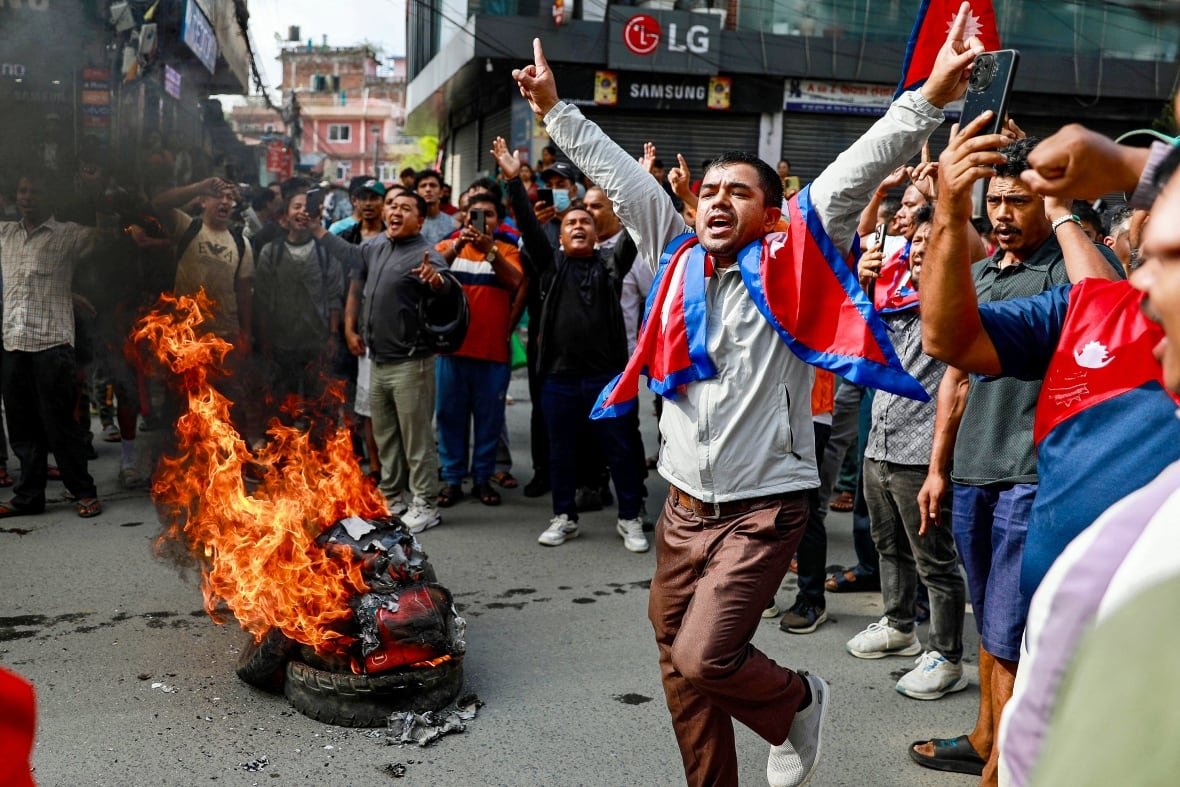 A man stands with a Nepalese flag on his neck and a burning tire can be seen.