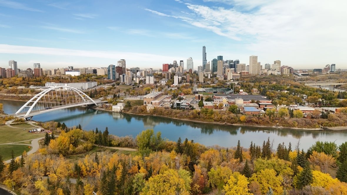 An aerial view of a downtown skyline with a river and a white bridge as leaves on the trees turn colour in the fall. 