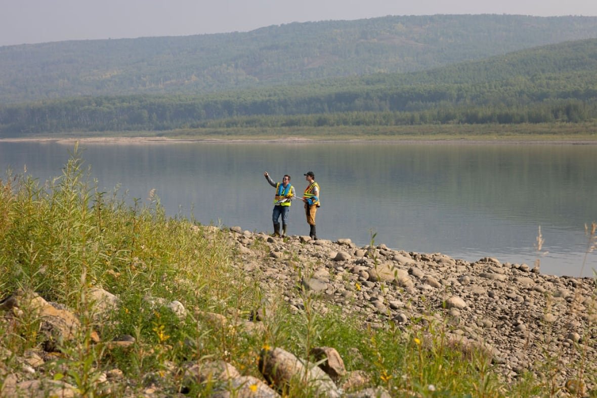 Two people in safety vests are pictured by a large body of water surrounded by pine trees.