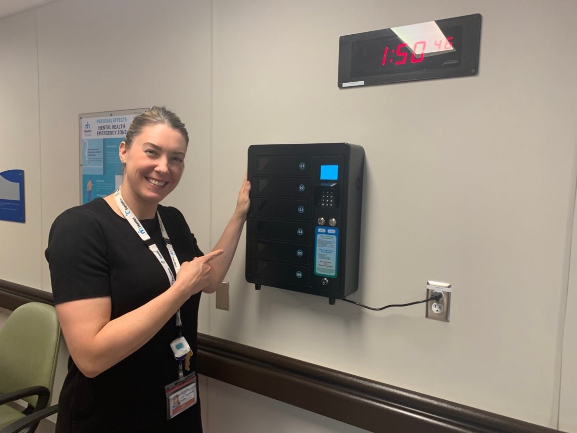 A woman with a ponytail smiles and points to a black box on the wall of a hospital waiting room.