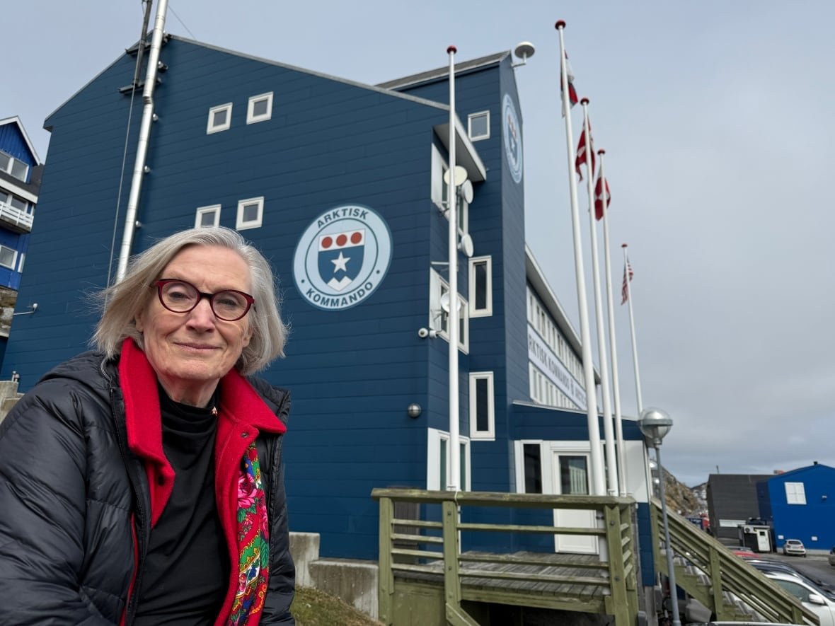 A woman with medium length grey hair smiles for a photo in front a blue building.