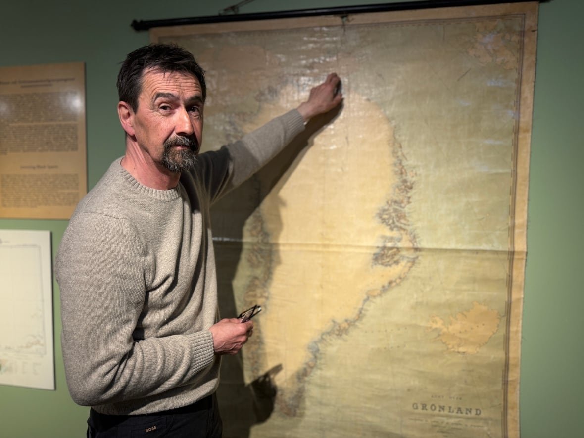 A man with shirt black hair wearing a grey sweater points to a map of Greenland. The map is faded and has a brown tint.