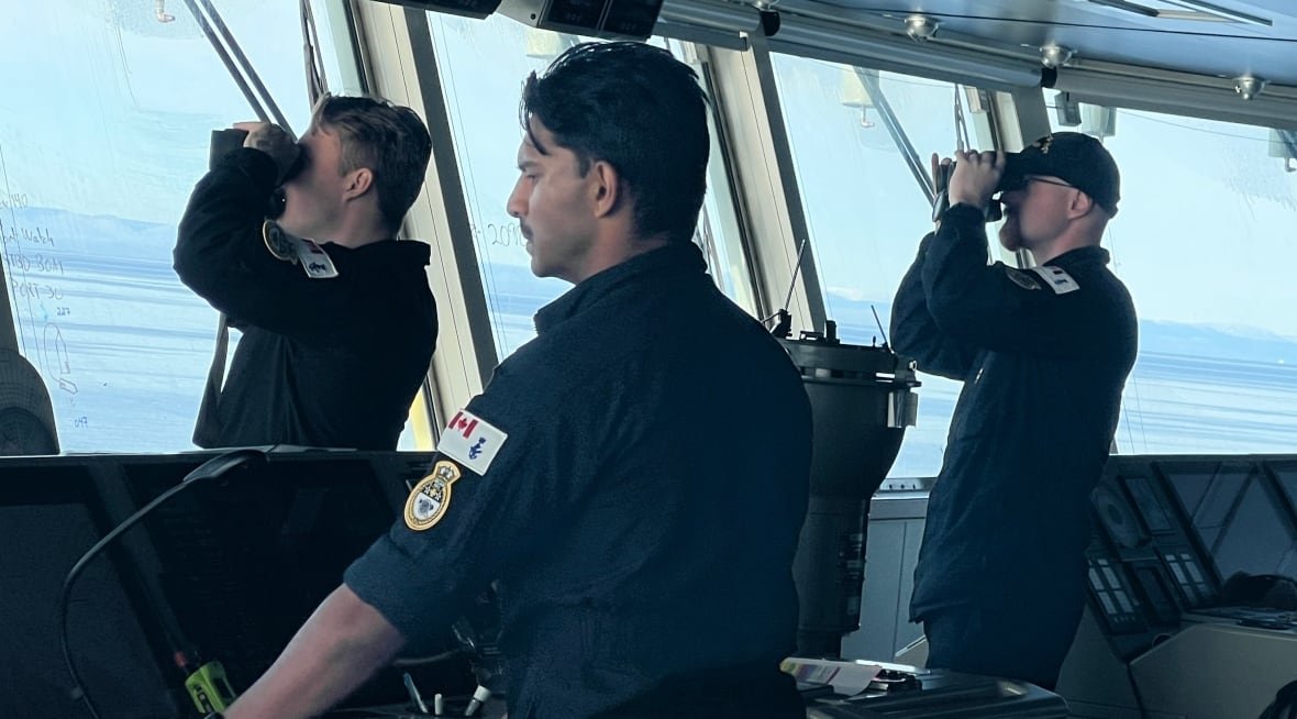 Three sailors stand on the bridge of a navy vessel -- one steering while the others peer through binoculars. 