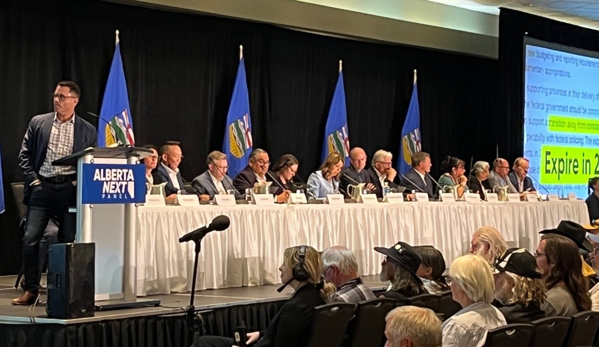 A panel of sixteen people, including Alberta premier Danielle Smith, sits at a long white table on a stage. Six Alberta flags hang on poles in the background.