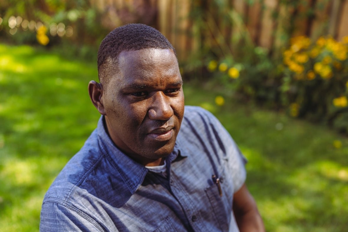 A Black man wearing a blue button up shirt sits in a backyard. 