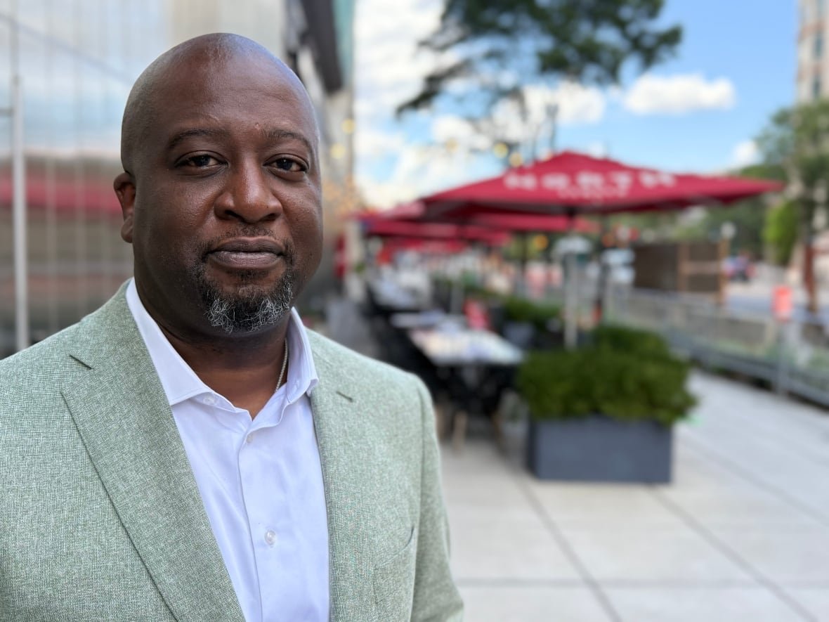 Shawn is an African American man with a bald head and a black and grey goatee. He is wearing a light grey suit with a white collared shirt with the first button open. He's standing in front of an empty patio with red umbellas at the tables.