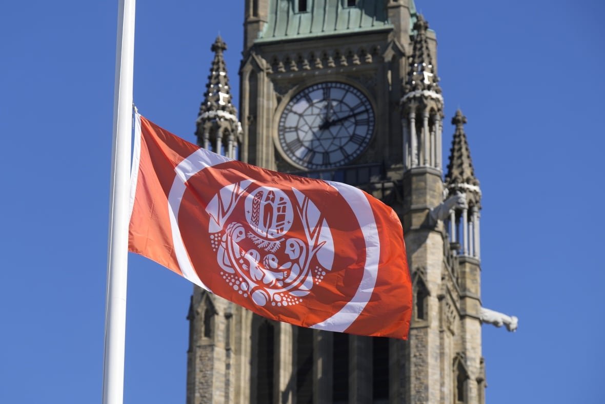 An orange and white flag with an Indigenous symbol waves in the wind in front of a clock tower.