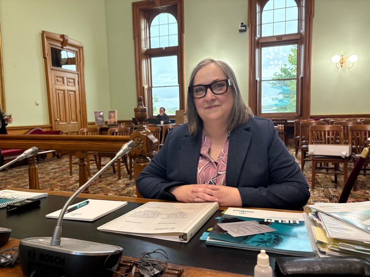 A woman with short grey hair wearing a navy blue blazer, black square-framed glasses, and a pink shirt sits at a desk in a legislative committee room with a large report sitting in front of her.